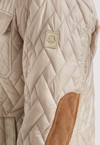 Beige gesteppte Jacke mit einem Cord-Elbogenflicken, einem Logo-Abzeichen auf dem Ärmel und einem diagonalen Steppmuster.