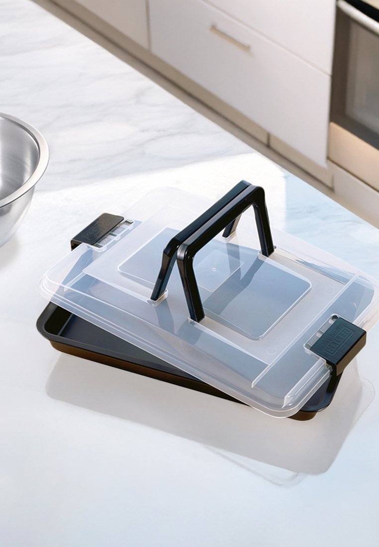 Transparente Kunststoff-Lebensmittelaufbewahrungsbox mit einem schwarzen Griff, stapelbarem Design, rechteckiger Form und schwarzem Boden. Silberne Rührschüssel in der Nähe.