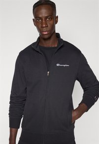 Μαύρο φούτερ με φερμουάρ, κατασκευασμένο από βαμβακερό μείγμα, με ψηλό γιακά, ριμπ μανσέτες και μικρό λευκό λογότυπο Champion στο αριστερό στήθος.
