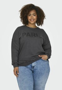 Šedá mikina s nápisem "PARIS" v černém písmu, kulatý výstřih, dlouhé rukávy a volný střih. Kombinováno s modrými džíny, neformální styl.