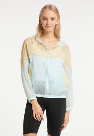Junge Frau trägt eine pastellfarbene Farb-blockierte Zip-Hoodie-Jacke und schwarze Radhosen, steht vor einem einfachen weißen Hintergrund.