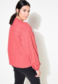Blusa corallo con un motivo floreale testurizzato, maniche lunghe e vestibilità ampia, abbinata a pantaloni neri. Il tessuto ha una leggera lucentezza.
