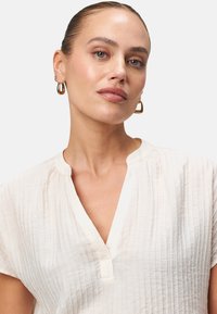 Blusa in crema leggera e strutturata con scollatura a V e maniche corte, caratterizzata da un design minimalista e sottili strisce verticali.