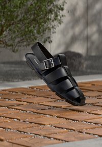 Μαύρο λαστιχένιο σανδάλι με κλειστή μύτη, λεπτομέρειες με λουράκια και κούμπωμα με αγκράφα, τοποθετημένο πάνω σε υφασμένο ξύλινο φόντο.