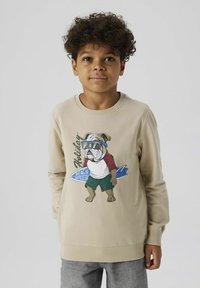 Sudadera beige con cuello redondo, que presenta un gráfico de un bulldog con gafas de sol. Mangas largas y puños acanalados, combinada con pantalones grises.