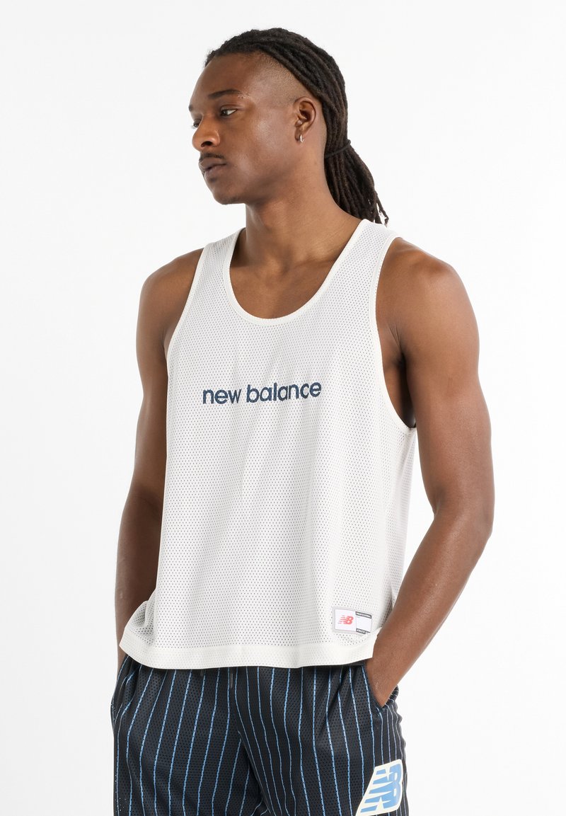 White mesh tank top with a loose fit, featuring "new balance" embroidered in navy blue. Paired with black and blue striped shorts.