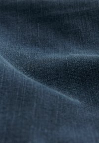 Denim tkanina u tamnoplavoj boji, s glatkom teksturom i suptilnim horizontalnim uzorkom. Materijal pokazuje blagi sjaj s mekanim naborima.