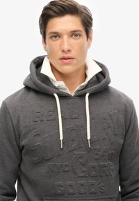 Sudadera gris oscuro con un estampado de logo texturizado, cordones blancos y un cuello blanco en contraste. Tejido suave con un ajuste relajado.