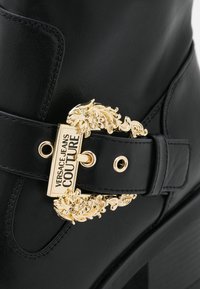 Botín de cuero negro con una hebilla decorativa dorada con detalles intrincados y una etiqueta rectangular que dice "VERSACE JEANS COUTURE."