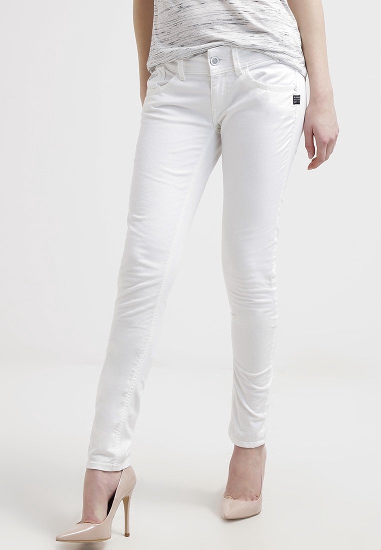 Femme portant un jean blanc slim et des chaussures à talons hauts couleur nude, debout avec une jambe légèrement pliée sur un fond neutre.