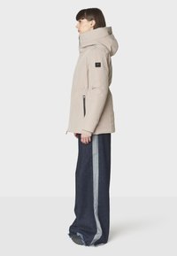 Chaqueta acolchada en beige claro, con capucha, dos bolsillos laterales con cremallera y cintura ajustada. Combinada con pantalones de mezclilla de pierna ancha con una raya gris.
