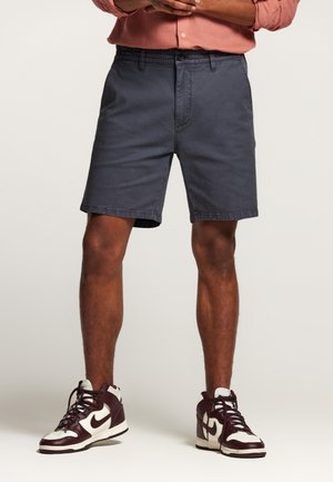 Shorts pour homme bleu marine avec fermeture à bouton, coupe droite et poches latérales. Associés à des baskets montantes bordeaux avec des accents blancs.