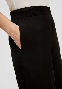 Zwarte broek met een elastische tailleband, voorzien van een zijzak en een gladde textuur. De stof is lichtgewicht met een subtiele glans.