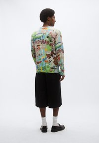 Ένα πολύχρωμο πουλόβερ με μακρύ μανίκι που διαθέτει ανάμεικτο γραφικό σχέδιο, κατασκευασμένο από ελαφρύ ύφασμα. Συνδυάζεται με μαύρο σορτς μέχρι το γόνατο και μαύρα παπούτσια.