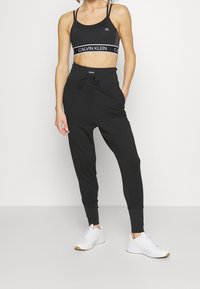 Kobieta ubrana w czarny sportowy biustonosz Calvin Klein, pasujące legginsy z wysokim stanem oraz białe sneakersy, stojąca na jednolitym tle.