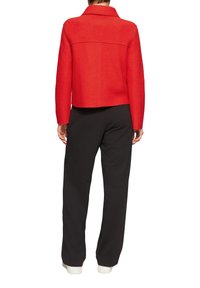 Czerwona krótka kurtka z mieszanki wełny, z kołnierzykiem z wycięciem i karczkiem z tyłu. Połączona z czarnymi spodniami i białymi ściągaczami.