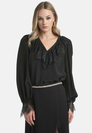 Black blouse featuring a V-neck and lace ruffle detail around the collar. Long sleeves with lace accents at the cuffs. Pleated black skirt.