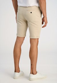 Beige shorts made of woven fabric, featuring rolled hems, back pockets with black buttons, worn with white sneakers. Minimalistic design.
