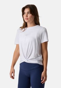 Femme aux cheveux bruns portant un haut blanc à manches courtes avec ourlet torsadé et des leggings bleu marine, debout devant un fond uni.