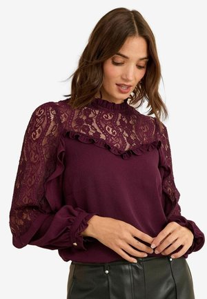 Vrouw draagt een paarse blouse met lange mouwen en kant op stukjes aan hals en mouwen, gecombineerd met zwarte leren broek, kijkt naar beneden met handen bij de taille.
