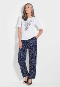Weißes T-Shirt mit schwarzem Blumenmuster und kurzen Ärmeln, kombiniert mit navy-gestreiften Hosen mit Punkten. Das Modell trägt offene Sandalen.