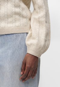 Main à la peau brune reposant sur le côté, portant un pull en maille crème avec manches bouffantes et un jean en denim bleu clair.