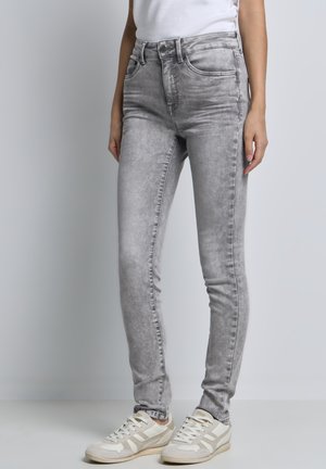 Persoon in grijze skinny jeans en witte sneakers, staand tegen een effen lichtgrijze achtergrond.