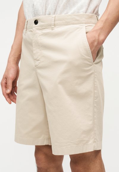 Άνδρας που φοράει μπεζ βερμούδα μέχρι το γόνατο με πλαϊνές τσέπες, έχει το ένα χέρι στην τσέπη, συνδυασμένη με ανοιχτόχρωμο πουκάμισο με υφή.