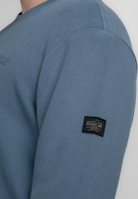 Blauwe gebreide sweatshirt met een ronde halslijn en een zwart label op de linkermouw met "PETROL IND" merknaam.