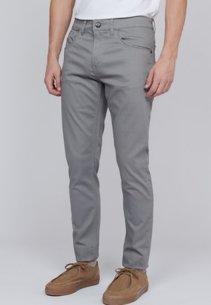 Man wearing slim-fit light gray pants with front pockets and tan suede lace-up shoes, standing against a neutral background.