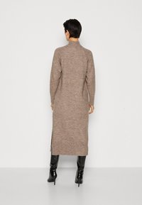 Robe longue en maille côtelée marron avec un col montant et des fentes latérales, associée à de longues bottes noires. Texture lisse, coupe ajustée.