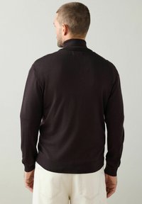 Mężczyzna odwrócony tyłem, ubraną w czarny sweter z długim rękawem z kołnierzykiem i białe spodnie, na jednolitym jasnym tle.