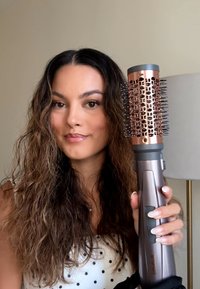 Vrouw met lang golvend haar die een ronde haarborstel met föhn dicht bij haar hoofd houdt, gekleed in een wit polkadot topje, neutrale achtergrond.