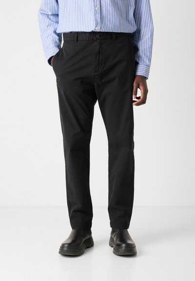 Pantalons en coton noir avec une coupe décontractée, dotés d'une fermeture par bouton à l'avant et de poches latérales, associés à des bottes noires et une chemise à rayures bleues.