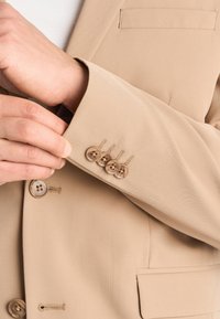 Hånd justerer manchet på beige blazer med en række af fire brune knapper og synlig lomme foran.