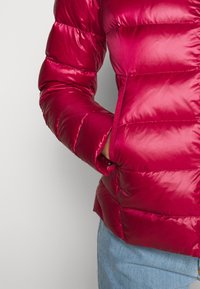 Veste matelassée rouge avec une texture brillante, présentant des sections matelassées. Comprend une poche latérale et une coupe ajustée. Portée avec un jean bleu.