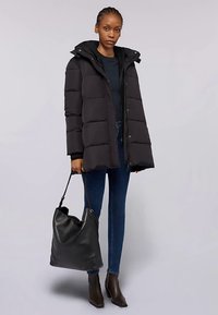 Svart vadderad jacka med huva, med horisontell sömnad, dragkedja och tryckknappsstängning, bärs med blå jeans och en svart läderväska.