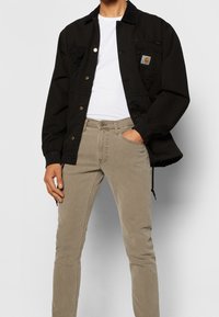 Svart kanvasjacka med knäppdesign, som har en framficka. Matchad med figurnära ljusbeige jeans. Enkel, funktionell stil.