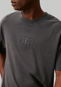 Záber zblízka na osobu v tmavě šedém tričku od Calvin Klein Jeans s vyšitým logem "CK" a textem na hrudi.