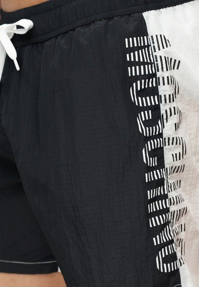 Μαύρες και άσπρες νάιλον σορτς με ελαστική μέση και λευκό κορδόνι, με κάθετο ριγέ κείμενο στην πλευρική επένδυση.