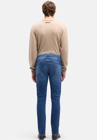 Beżowa koszula z długim rękawem z metką z tyłu, w zestawie z niebieskimi dżinsami z denimu. Dżinsy mają dopasowany krój i widoczne tylne kieszenie.
