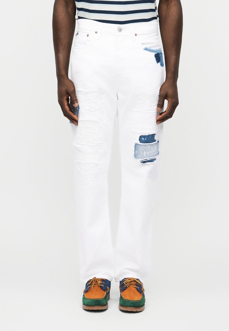 Άτομο που φοράει άσπρο τζιν με φθορές και μπλε μπαλώματα, ριγέ πουκάμισο μέσα από το παντελόνι και πολύχρωμα παπούτσια με κορδόνια σε πολλαπλούς τόνους.