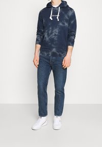 Námořnická batikovaná mikina s bílými šňůrkami, přední kapsou a logem, kombinovaná s modrými džínami a bílými teniskami.