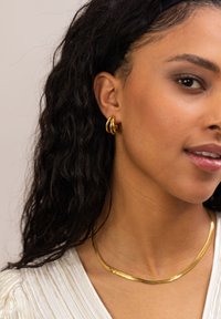 Goldene Ohrhänger und eine gewellte goldene Halskette an einem Model. Strukturierter weißer Oberteil; weiche Locken umrahmen das Gesicht. Minimalistisches Design, elegante Merkmale.