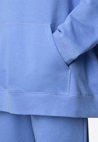 Sweat-shirt bleu clair en tissu doux, doté d'une poche kangourou et de poignets côtelés. Détail de couture visible le long des coutures.