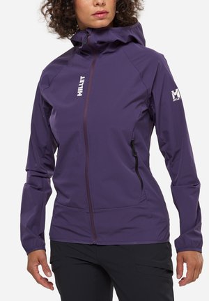Femme portant une veste à capuche ajustée de couleur violette avec des poches zippées à l'avant et sur les côtés, associée à un pantalon noir, sur un fond blanc.