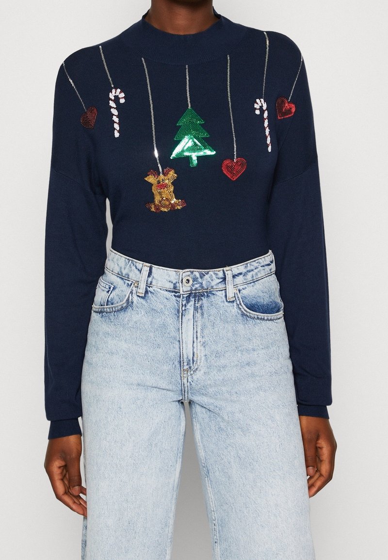 Pull bleu marine avec décorations de Noël en sequins suspendues, comprenant des cannes de bonbon, des cœurs, un sapin et un renne, associé à un jean bleu clair.