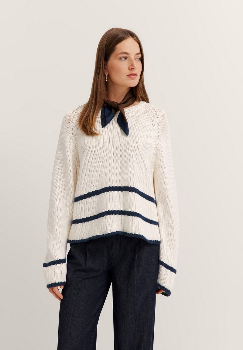 Cremefarbener Pullover mit marineblauen Streifen am Saum und an den Bündchen. Strukturiertes Strickmuster, Crew-Halsausschnitt. Kombiniert mit dunklen Jeans und einem braunen Halstuch.