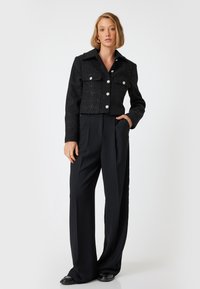 Veste en tweed noir coupée avec des boutons argentés, grand col et deux poches avant, assortie à un pantalon noir taille haute et des chaussures plates.