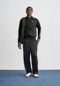 Juodas sportinis švarkas su baltais dryžiais ir logotipu, derinamas su atitinkančiomis kelnėmis. Balti sportbačiai užbaigia aprangą. Fonas yra paprastas.